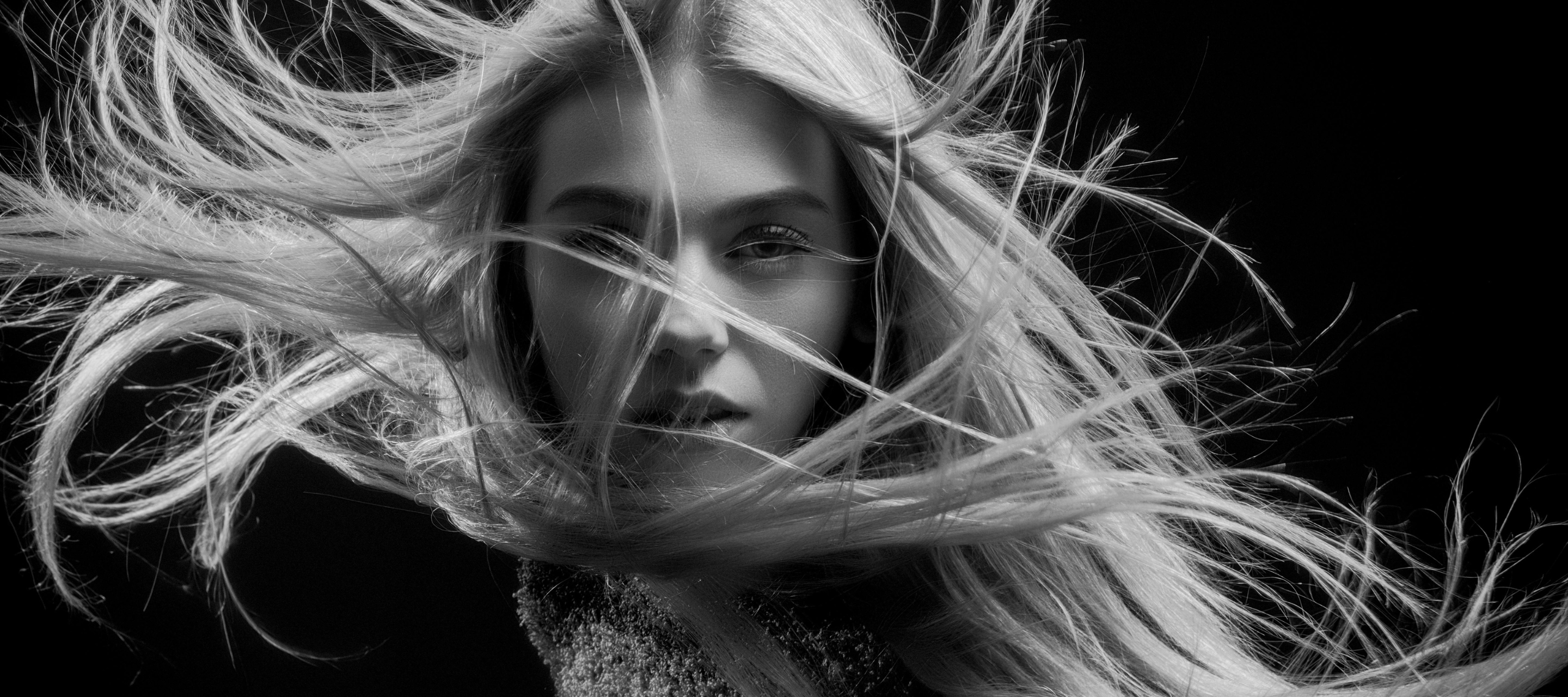 Black-and-white close-up portrait of a woman with long blonde hair blowing across her face, gazing directly at the camera