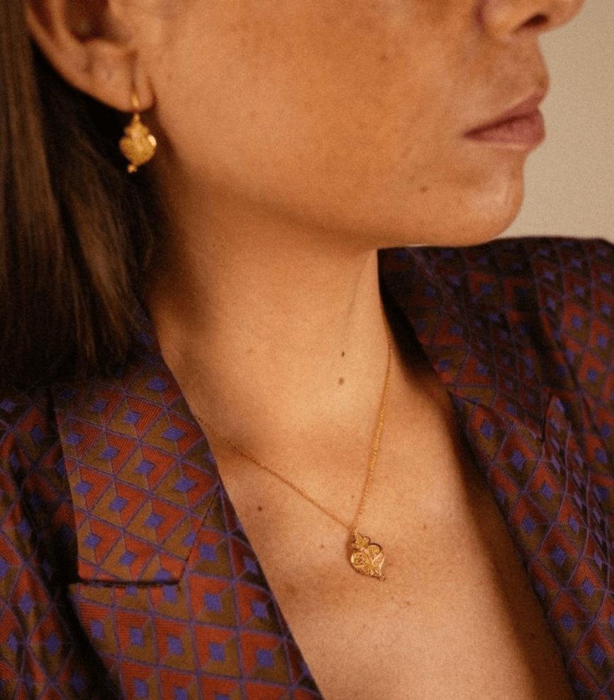 Close-up of a woman’s jawline and neck wearing a gold pendant necklace and matching gold earrings, with a patterned blazer in warm tones